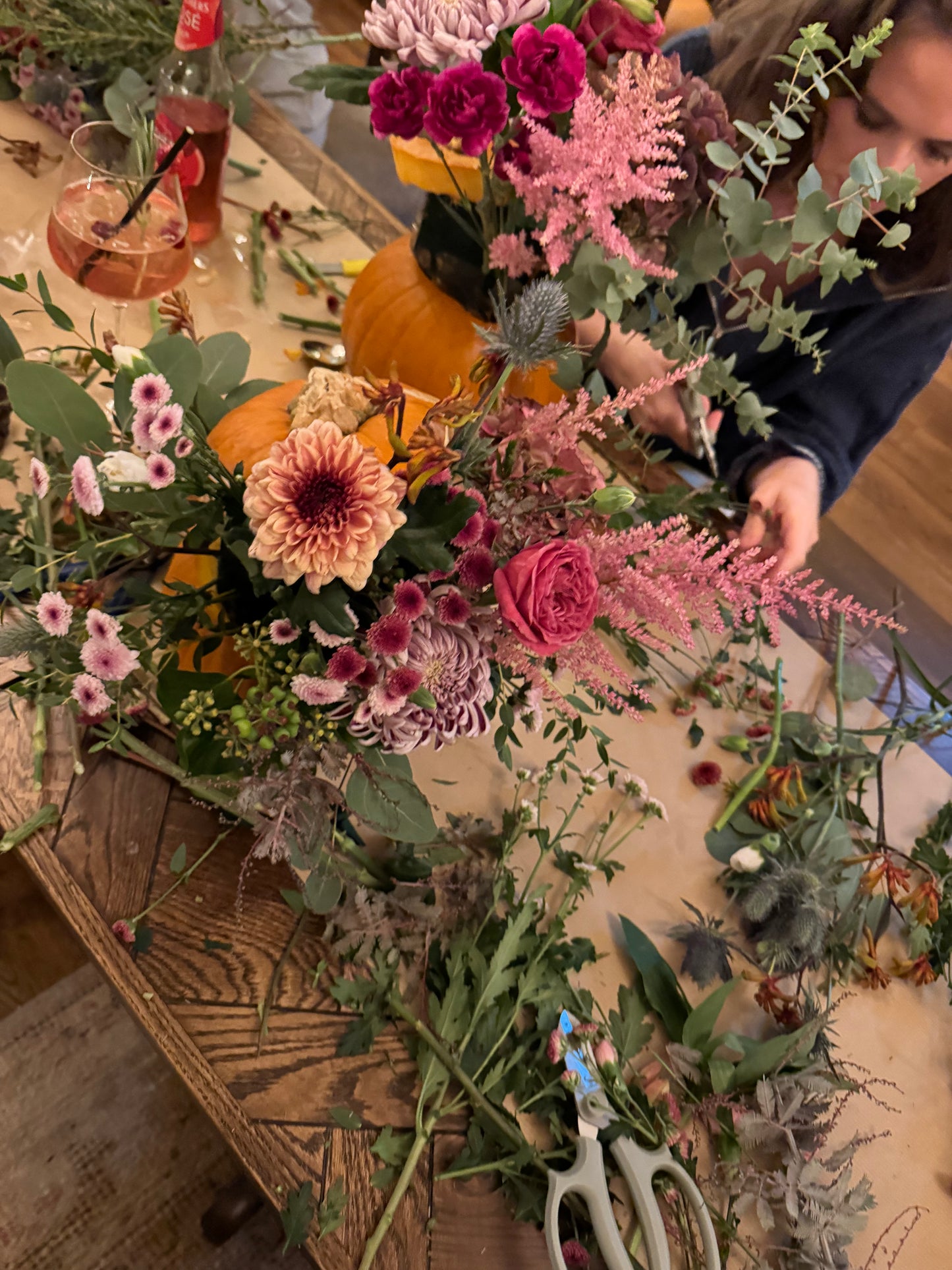 A close-up, overhead view of a florist arranging a large, colorful floral arrangement on a rustic wooden table. The bouquet features pink astilbe, burgundy roses, purple thistles, and orange dahlias, with pumpkins and floral scissors nearby.