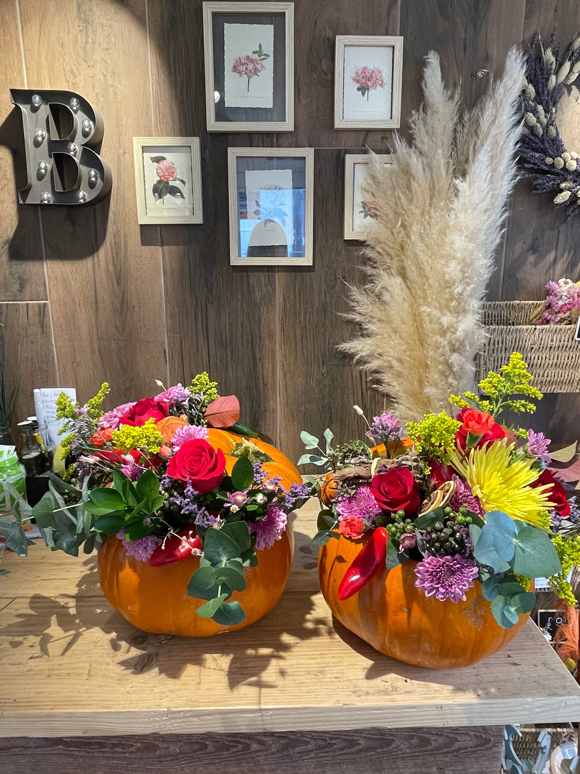Two orange pumpkins filled with colorful autumn floral arrangements, displayed on a wooden table against a wood-paneled wall with framed botanical prints.